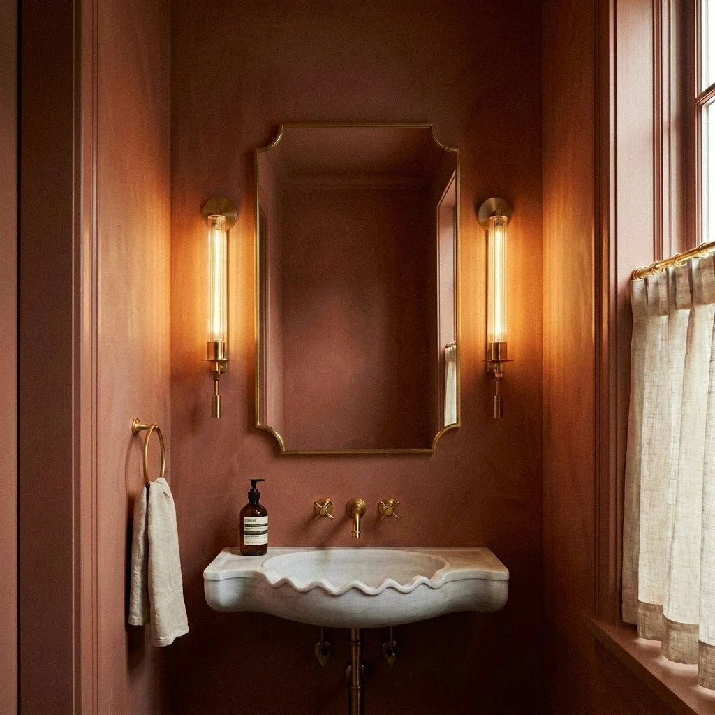 Pair of Clérachic gold wall lights with fluted glass tubes flanking a brass-framed mirror above a marble basin in a rust-pink room.