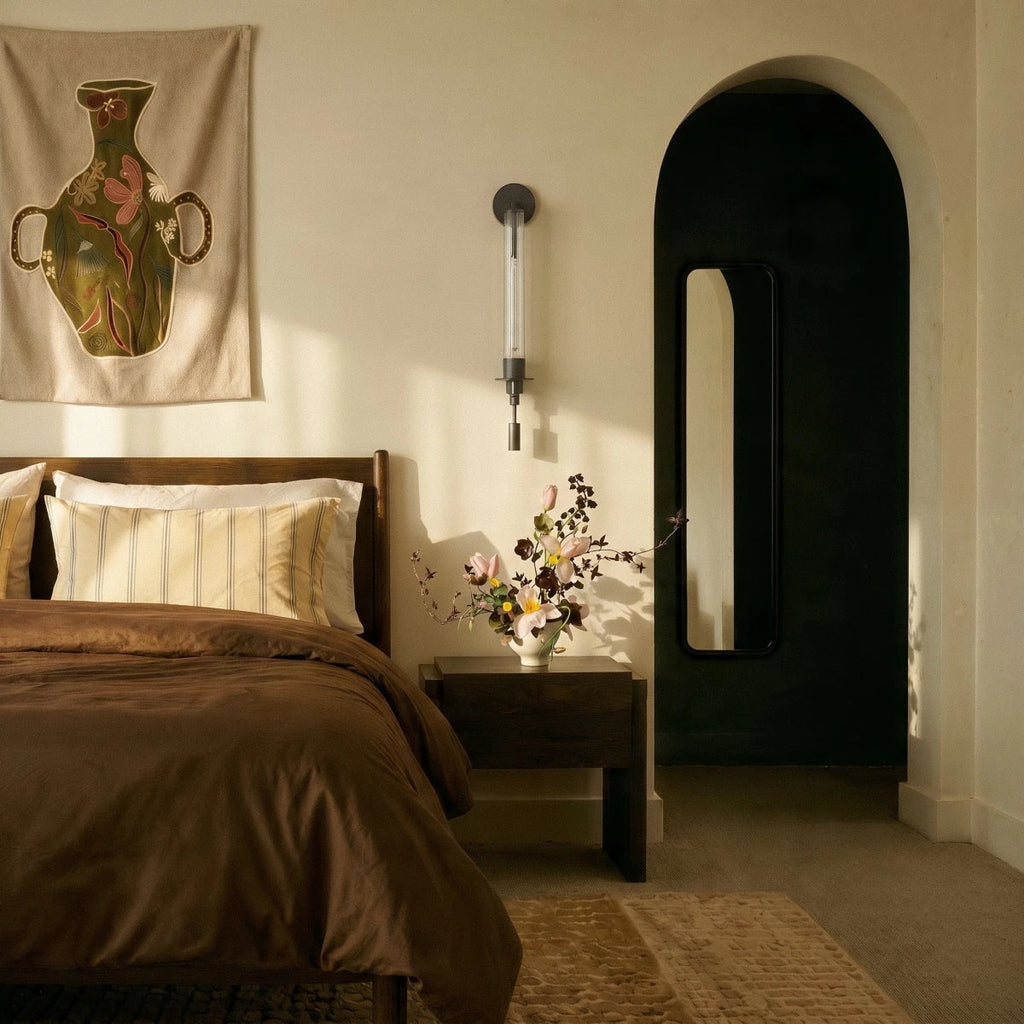 Clérachic black wall light with a fluted glass tube mounted beside a bed, with a brown duvet and an arched doorway nearby.
