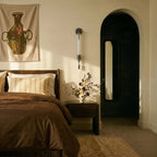Clérachic black wall light with a fluted glass tube mounted beside a bed, with a brown duvet and an arched doorway nearby.