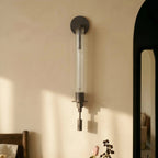 Clérachic black wall light with a round backplate and fluted clear glass tube, mounted on a cream wall in raking sunlight.