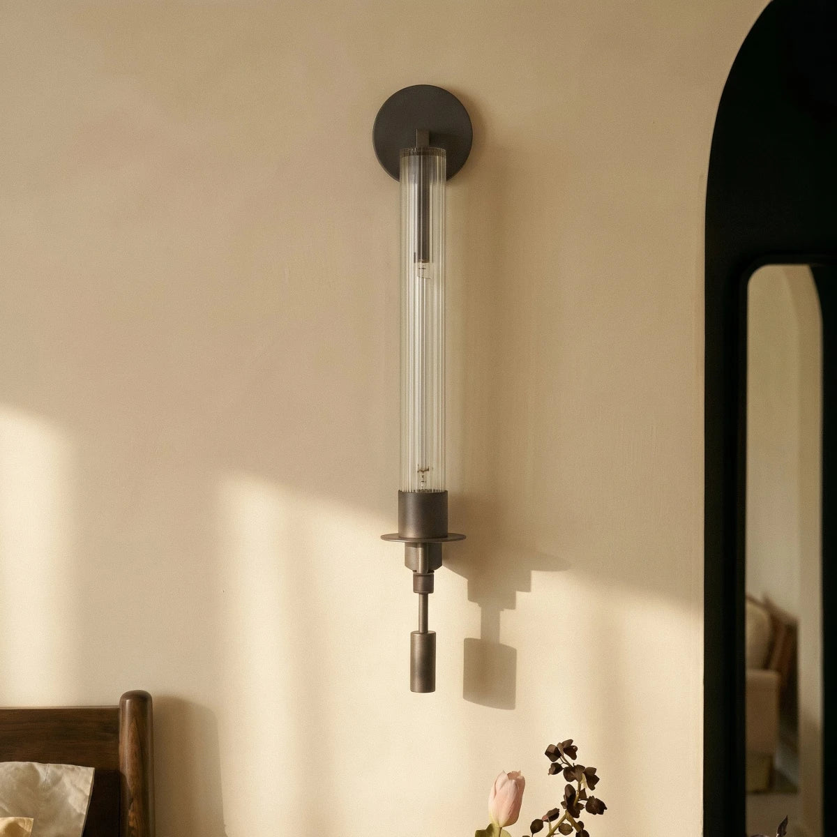 Clérachic black wall light with a round backplate and fluted clear glass tube, mounted on a cream wall in raking sunlight.