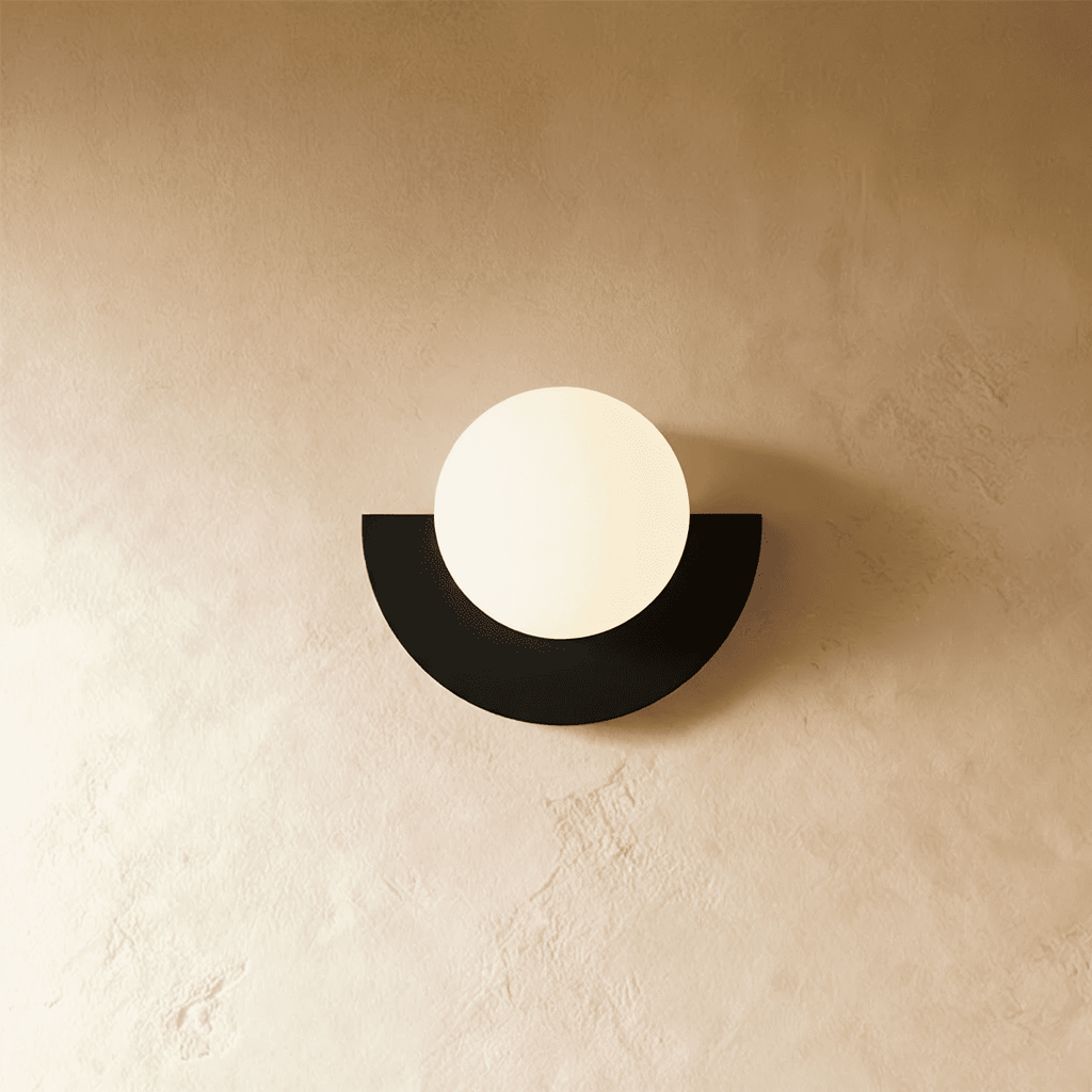 Clérachic lit opal glass globe on a black half-moon metal back plate, glowing softly on a limewash plaster wall.