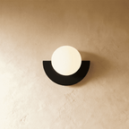 Clérachic lit opal glass globe on a black half-moon metal back plate, glowing softly on a limewash plaster wall.