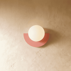 Clérachic lit opal glass globe on a coral-pink half-moon metal back plate, glowing softly on limewash plaster.