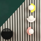 Clérachic globe wall lamps with yellow, white and pink half-moon plates on a striped wall, beside a black slatted round fixture.