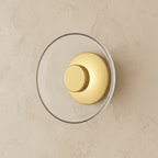 Clérachic glass uplight wall bowl in Transparent, a clear glass dome over a brass centre knob, unlit on a plain wall.