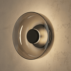 Clérachic glass uplight wall bowl in Smoky gray, its tinted dome glowing warm around a black centre knob.