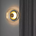Clérachic glass uplight wall bowl in Amber glows warm beside a dark curtain, its dome casting soft light upward.