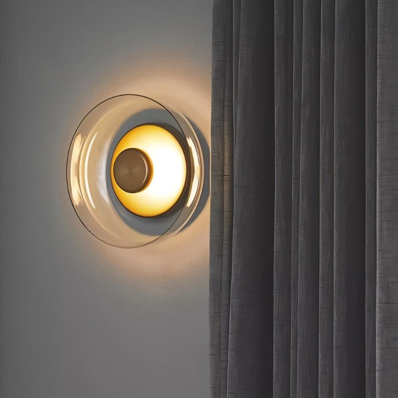 Clérachic glass uplight wall bowl in Amber glows warm beside a dark curtain, its dome casting soft light upward.