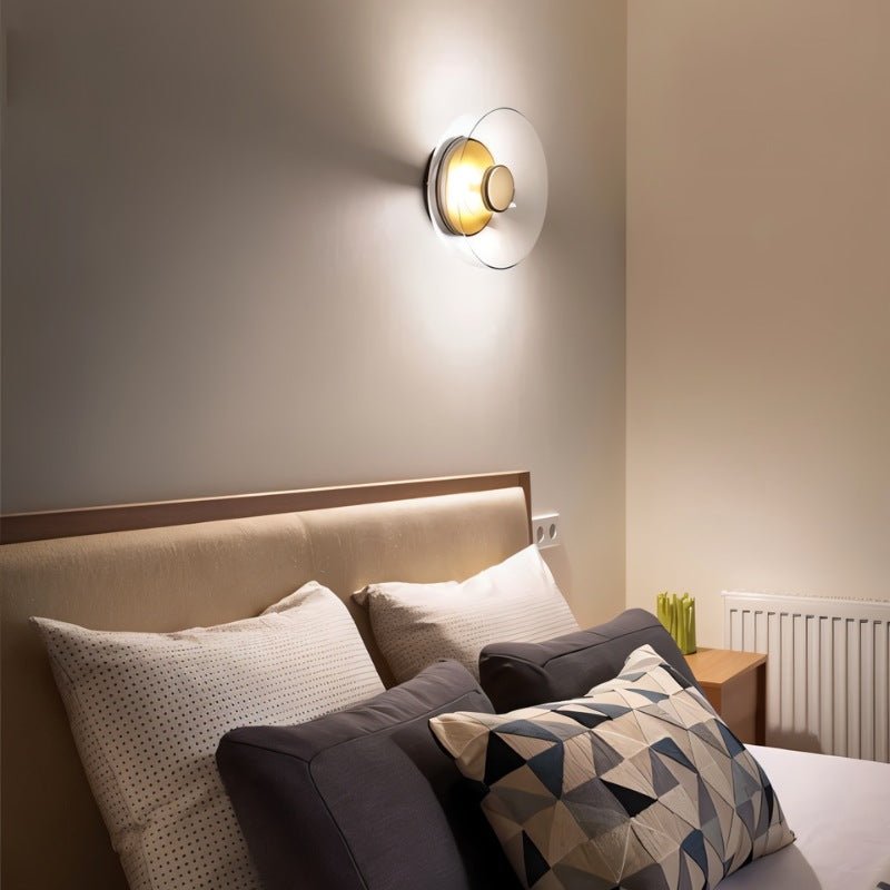 Clérachic glass uplight wall bowl glows warm above a bed, casting soft upward light across a plaster bedroom wall.