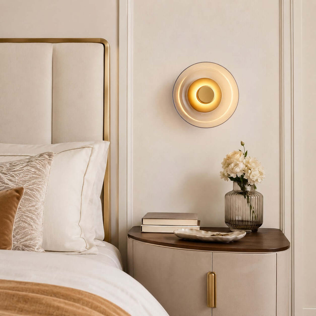 Lit Clérachic glass wall light on a cream panelled wall above a curved bedside cabinet, with white flowers and a padded headboard.