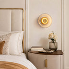 Lit Clérachic glass wall light on a cream panelled wall above a curved bedside cabinet, with white flowers and a padded headboard.
