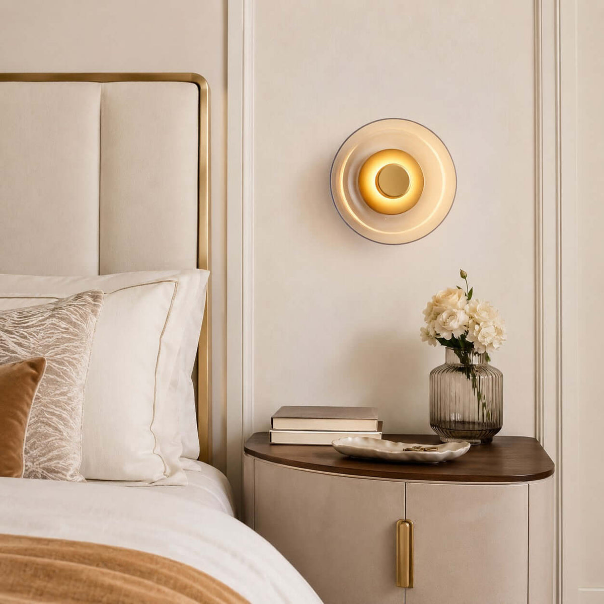 Lit Clérachic glass wall light on a cream panelled wall above a curved bedside cabinet, with white flowers and a padded headboard.