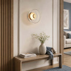 Clérachic round wall light glowing in a gold-and-clear finish above a wooden bench in a hall with slatted wood panelling.