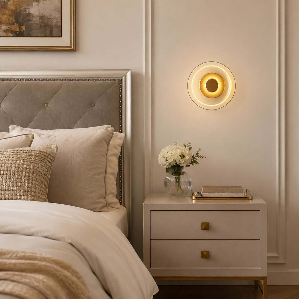 Lit Clérachic wall light above a cream two-drawer bedside table, with a button-tufted headboard and a small vase of white flowers.