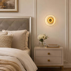 Lit Clérachic wall light above a cream two-drawer bedside table, with a button-tufted headboard and a small vase of white flowers.