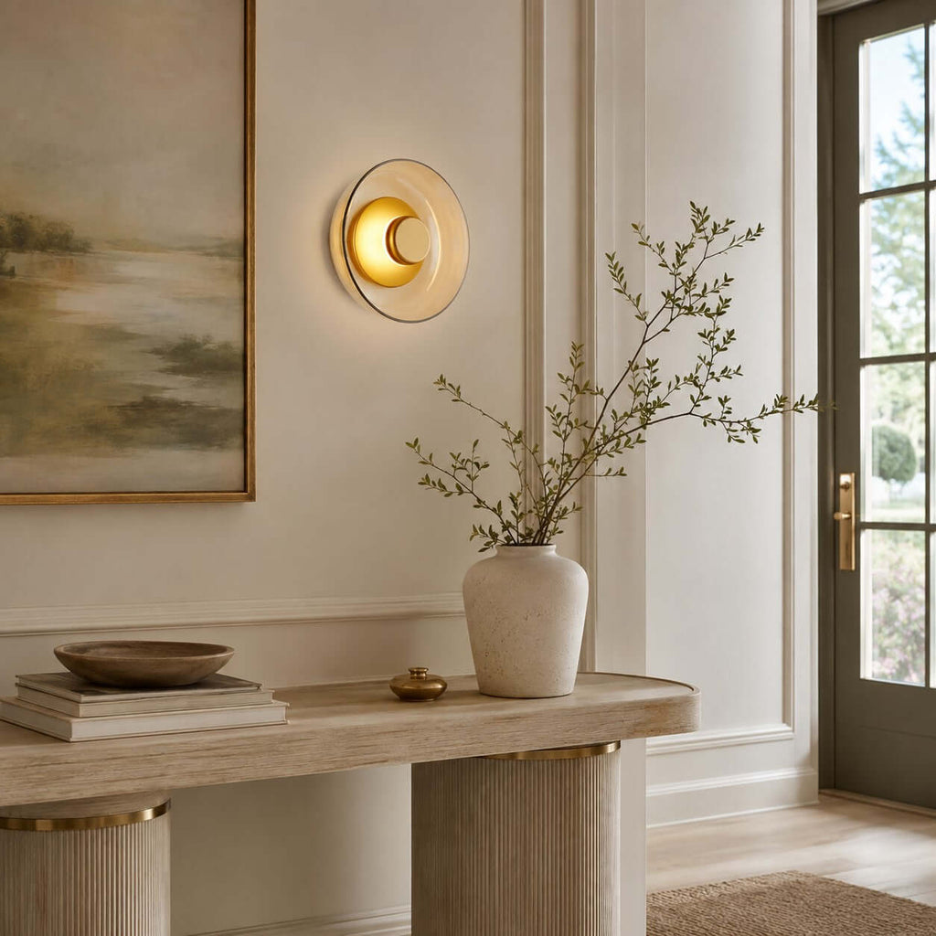 Clérachic round glass wall light glowing on a panelled wall, over a pale console with stacked books and a vase of bare branches.