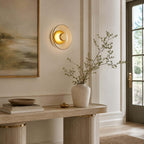 Clérachic round glass wall light glowing on a panelled wall, over a pale console with stacked books and a vase of bare branches.
