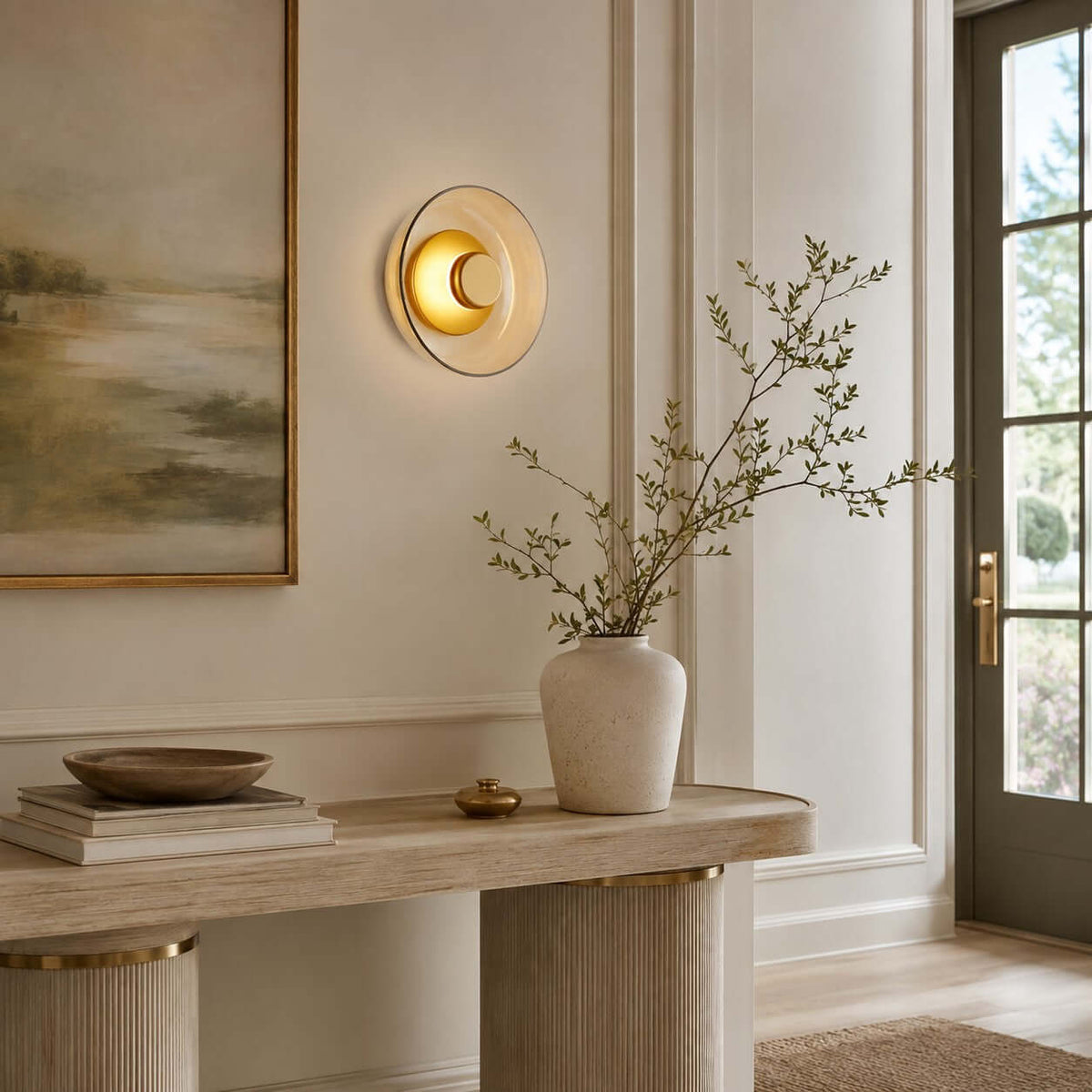 Clérachic round glass wall light glowing on a panelled wall, over a pale console with stacked books and a vase of bare branches.