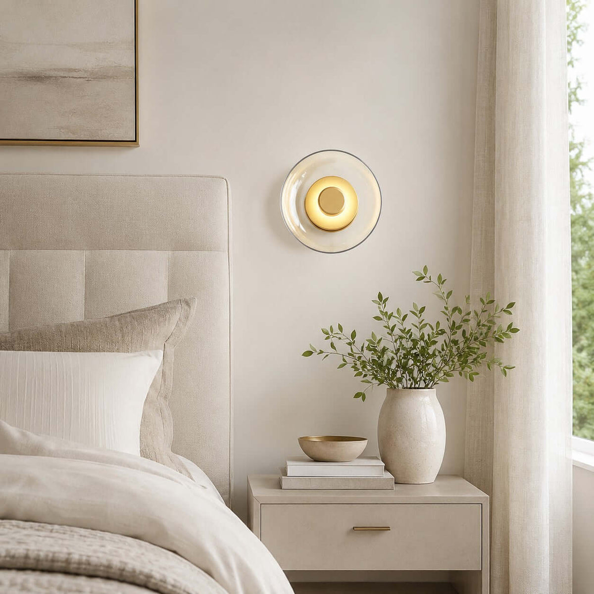 Clérachic glass wall light lit above a white bedside table, with a linen headboard, a cream vase of leaves and a sheer curtain.