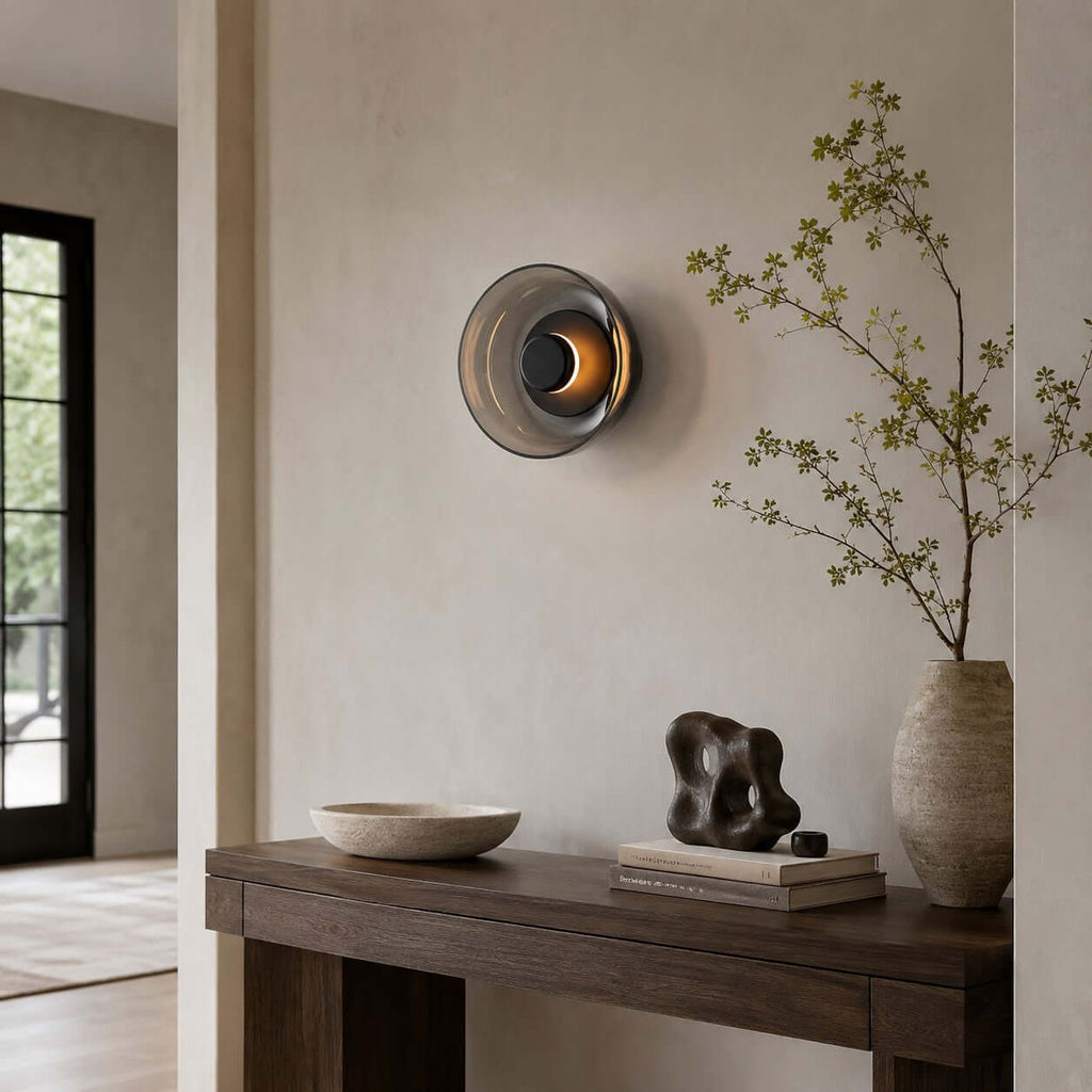Smoky grey version of this Clérachic wall light glowing amber above a dark wood console with a sculpture and a bowl.