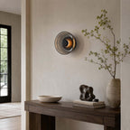 Smoky grey version of this Clérachic wall light glowing amber above a dark wood console with a sculpture and a bowl.
