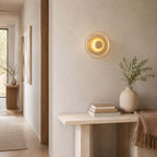 Clérachic wall light lit on a warm plaster wall in a corridor, above a pale console with books, a throw and a vase of olive leaves.