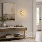 Clérachic glass wall light lit beside a large framed landscape, above a marble-topped console with books and a stone bowl.