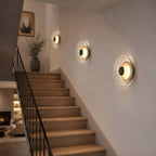 Three Clérachic wall lights glowing in a row up a staircase wall, beside a black railing and a vase of branches.