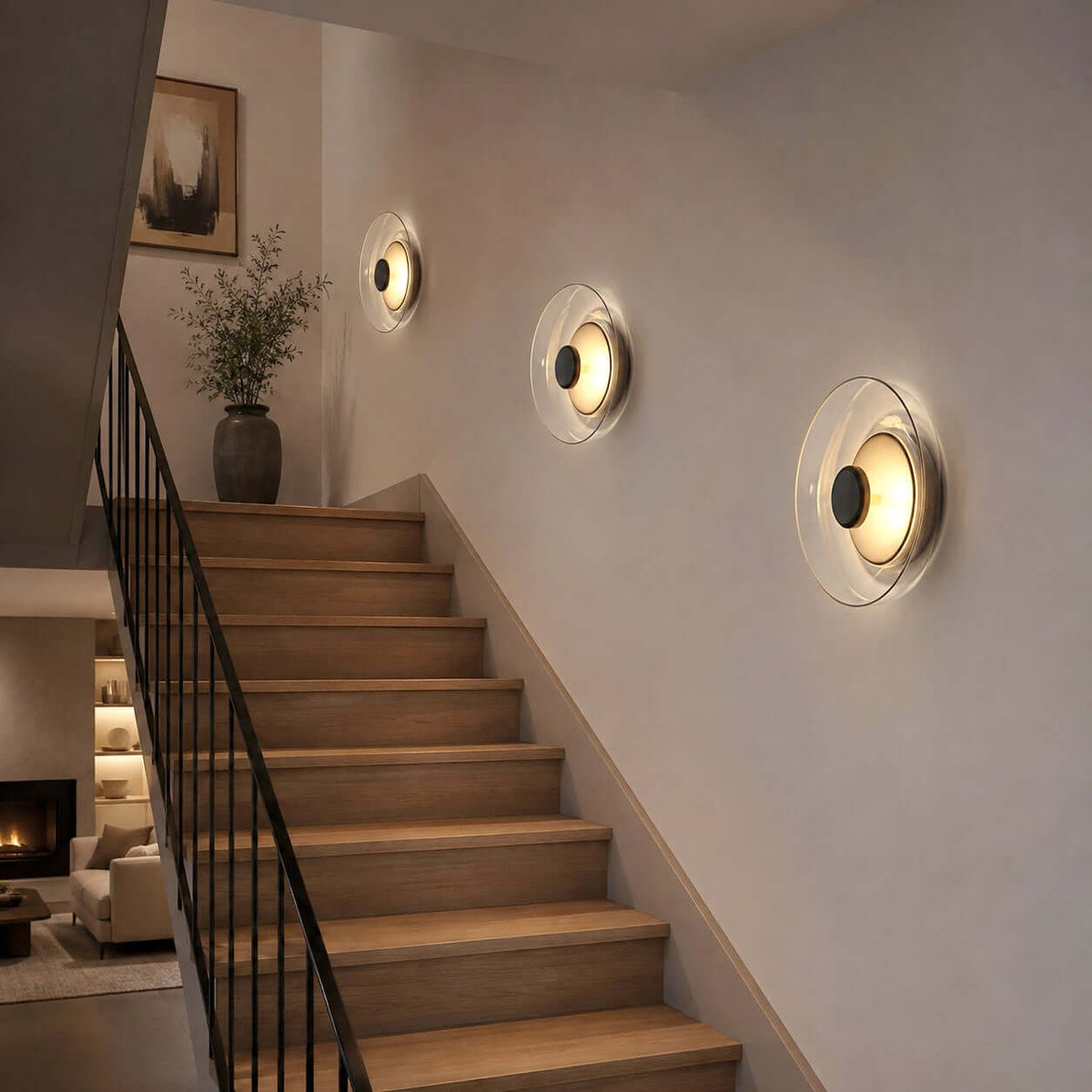 Three Clérachic wall lights glowing in a row up a staircase wall, beside a black railing and a vase of branches.