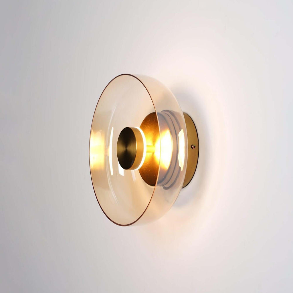 Clérachic wall light lit on a pale wall, an amber-tinted glass disc around a gold centre cap and a glowing bulb.