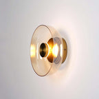 Clérachic wall light lit on a pale wall, an amber-tinted glass disc around a gold centre cap and a glowing bulb.