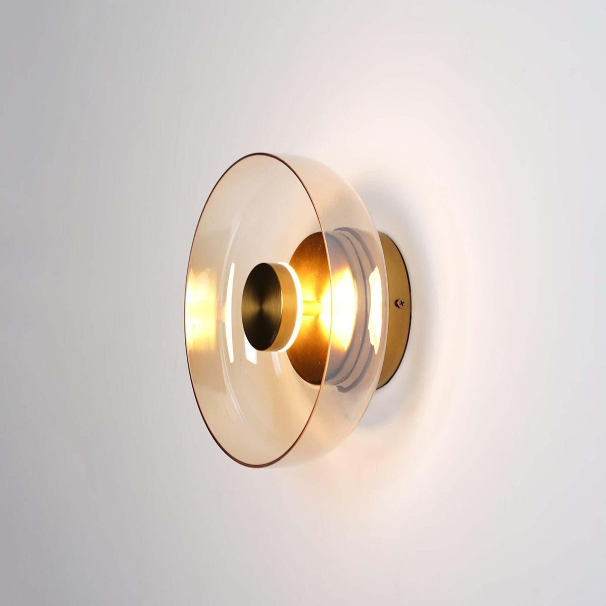Clérachic wall light lit on a pale wall, an amber-tinted glass disc around a gold centre cap and a glowing bulb.