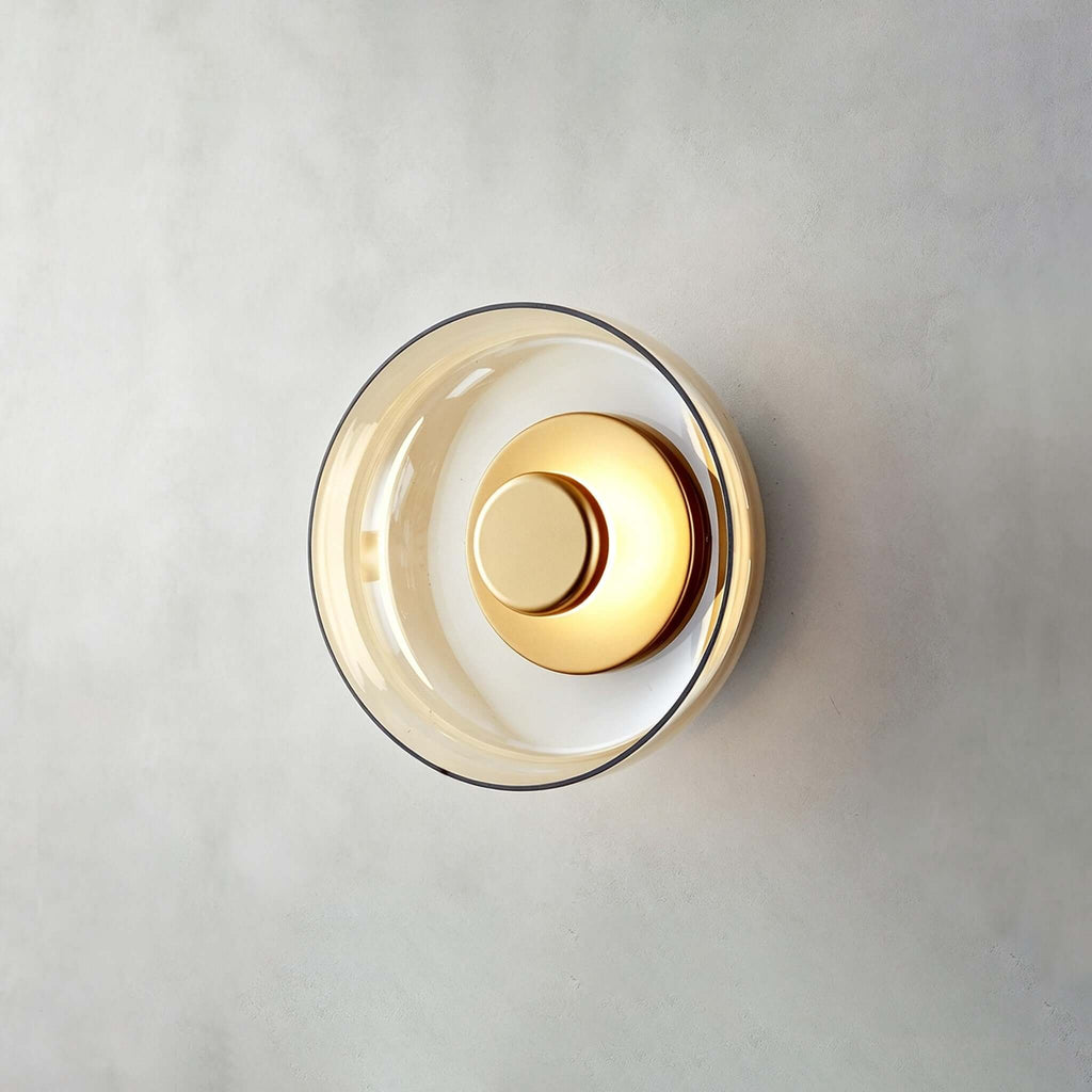 Front view of a Clérachic wall light in gold and amber on a grey plaster wall, the glass disc edged in warm tint.