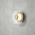 Clérachic wall light in gold and clear on grey plaster, a transparent glass disc around a pale gold centre cap.