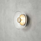 Clérachic wall light in gold and clear on grey plaster, a transparent glass disc around a pale gold centre cap.