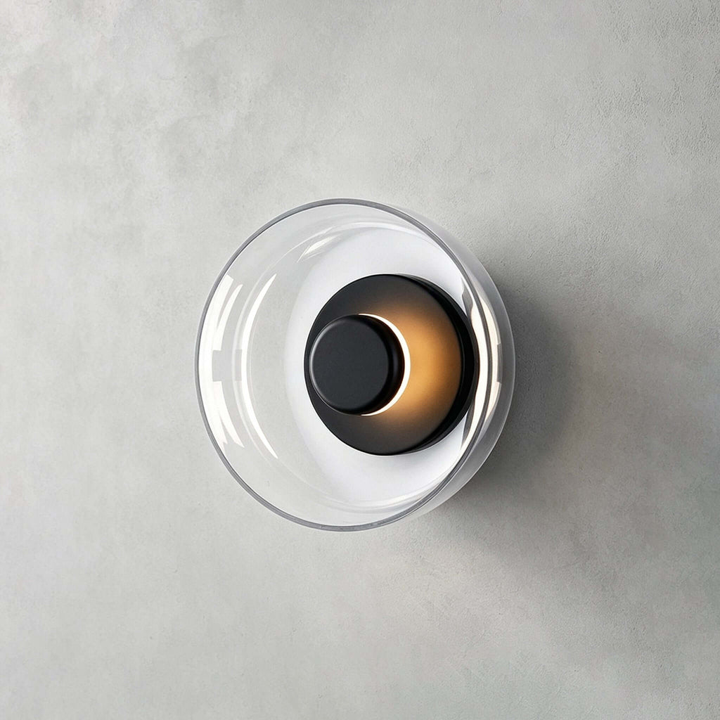 Clérachic wall light in black and clear glass on grey plaster, with a dark centre cap and a warm glow at its edge.