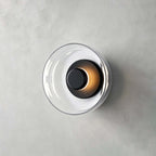 Clérachic wall light in black and clear glass on grey plaster, with a dark centre cap and a warm glow at its edge.