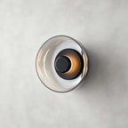 Clérachic wall light in black and smoky grey on a concrete-effect wall, the dark glass disc holding a glowing black cap.
