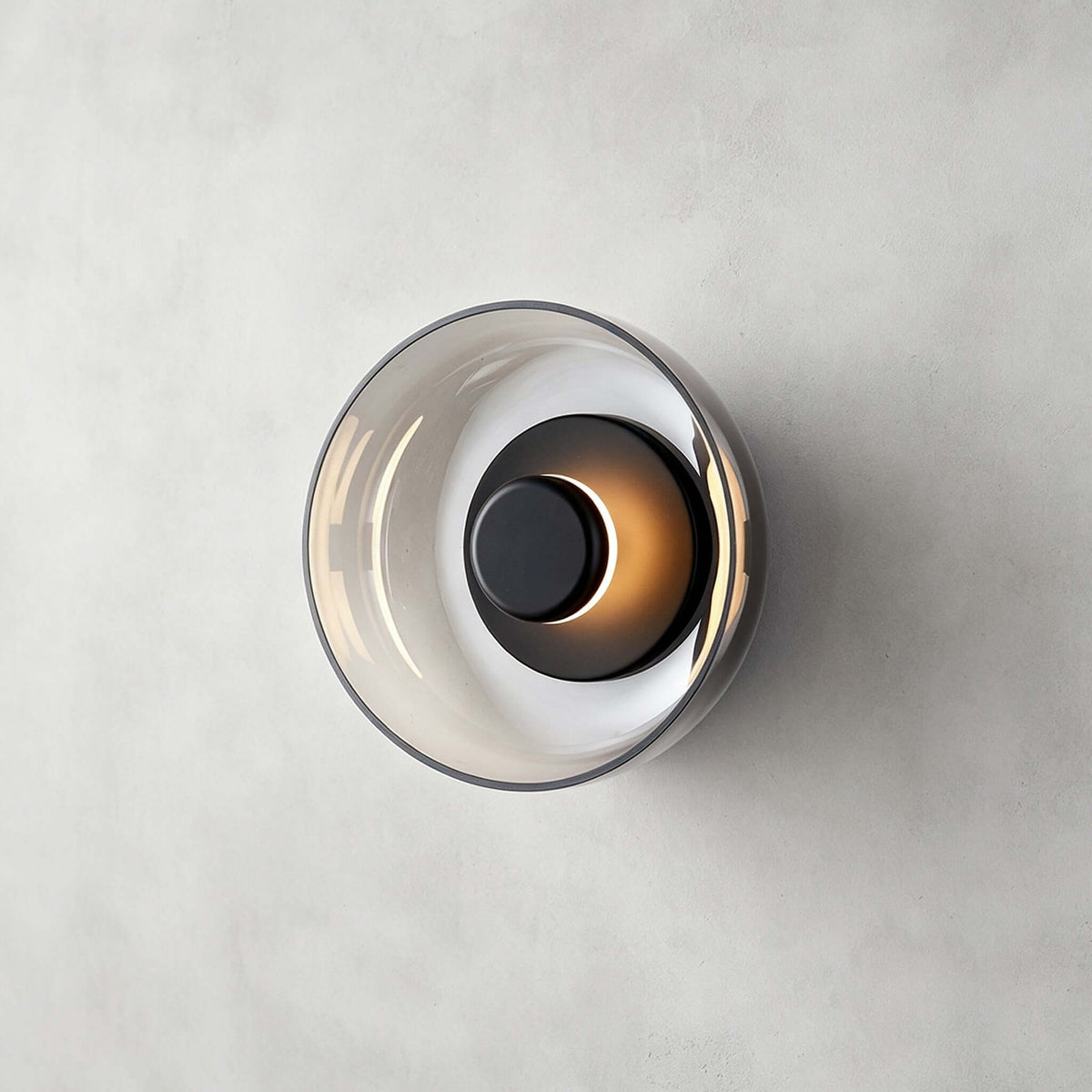 Clérachic wall light in black and smoky grey on a concrete-effect wall, the dark glass disc holding a glowing black cap.