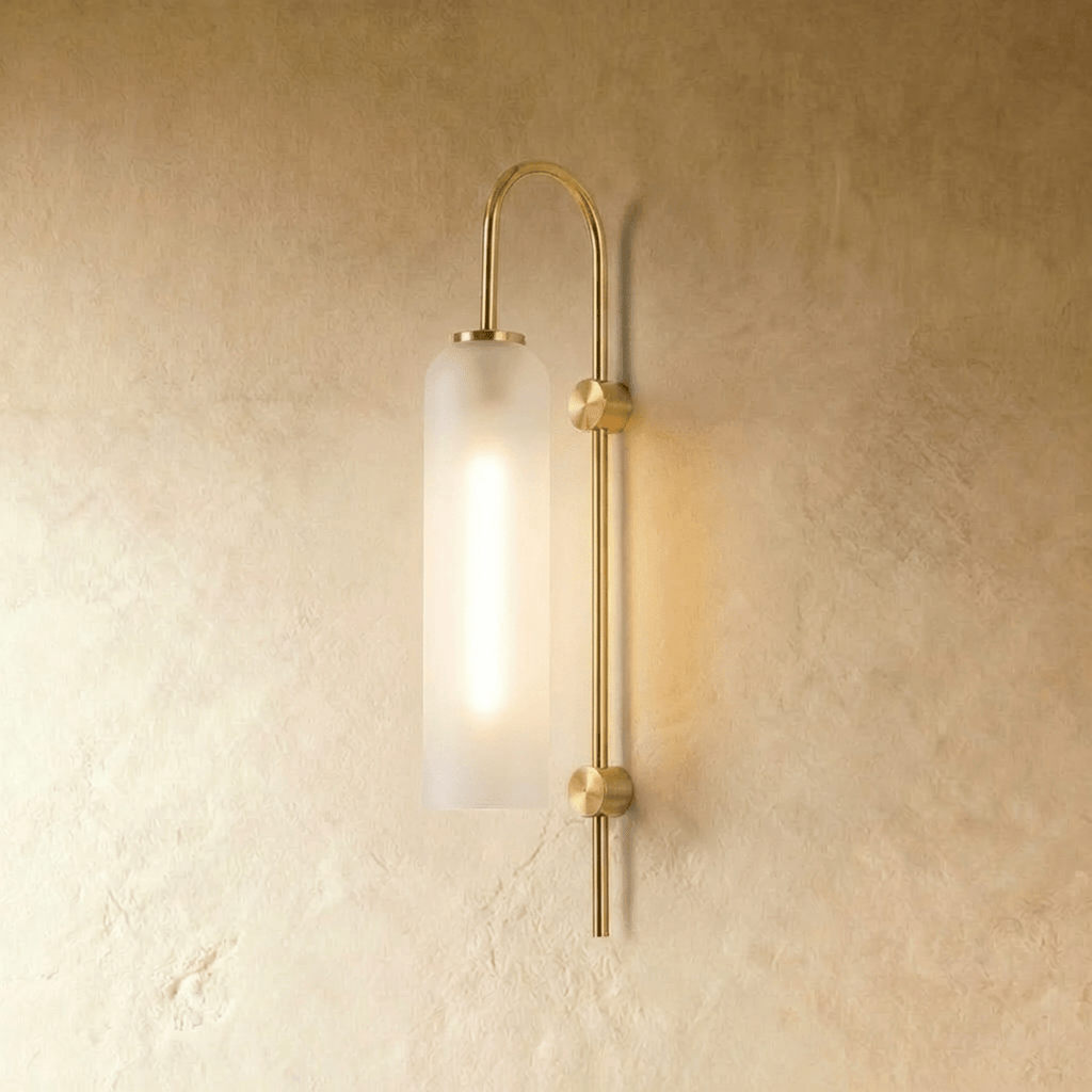 Clérachic wall light with a tall frosted glass cylinder on an arched brass arm, glowing softly against a limewashed wall.