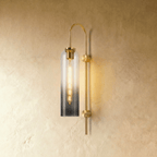Clérachic wall light with a smoky gradient glass cylinder on an arched brass arm, a long filament bulb glowing inside.