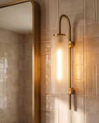 Close view of a Clérachic brass wall light with a frosted glass cylinder, lit, on glossy cream tiles beside a brass mirror frame.