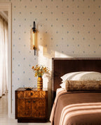 Clérachic black wall light with an amber glass cylinder, lit above a burl wood bedside table in a bedroom with floral wallpaper.