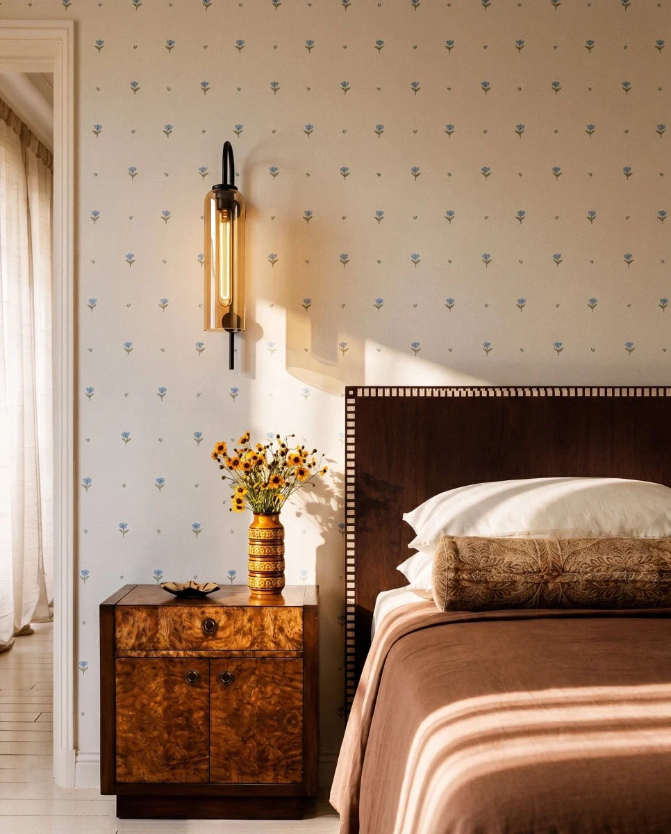 Clérachic black wall light with an amber glass cylinder, lit above a burl wood bedside table in a bedroom with floral wallpaper.