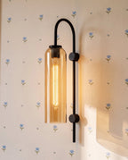 Close view of a Clérachic black arched wall light with an amber glass cylinder, its tube bulb glowing on floral wallpaper.