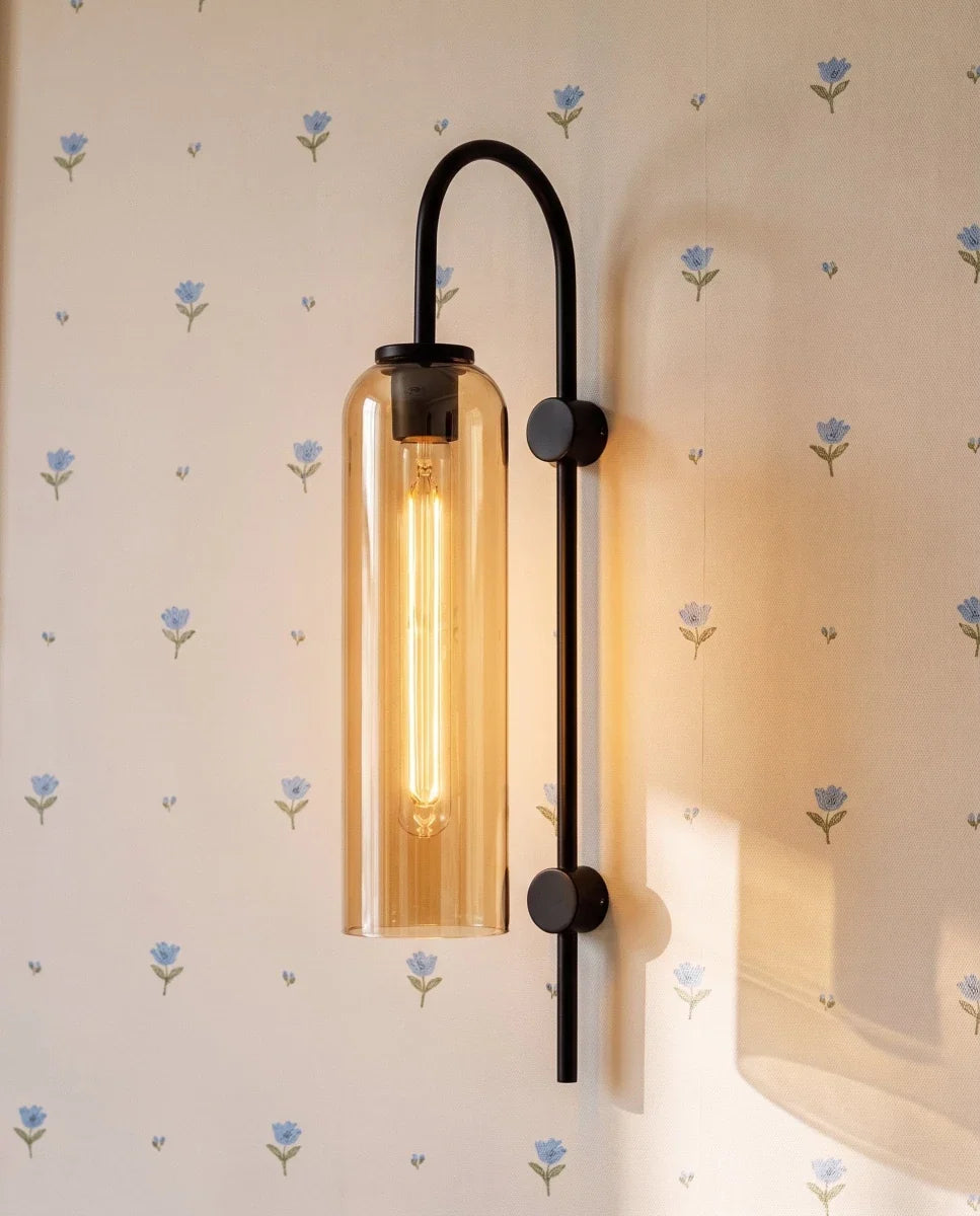 Close view of a Clérachic black arched wall light with an amber glass cylinder, its tube bulb glowing on floral wallpaper.