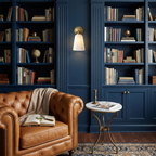One gold Clérachic wall sconce glows between navy blue bookshelves, above a tan leather armchair and a small marble side table.