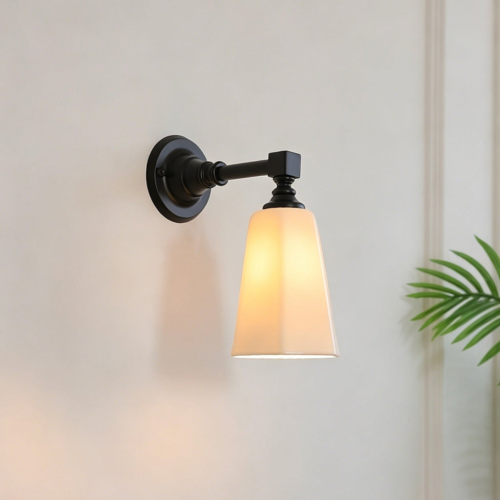 Lit black and white Clérachic wall sconce with a tapered glass shade, against a pale plaster wall with a palm leaf at the edge.
