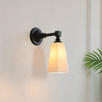 Lit black and white Clérachic wall sconce with a tapered glass shade, against a pale plaster wall with a palm leaf at the edge.
